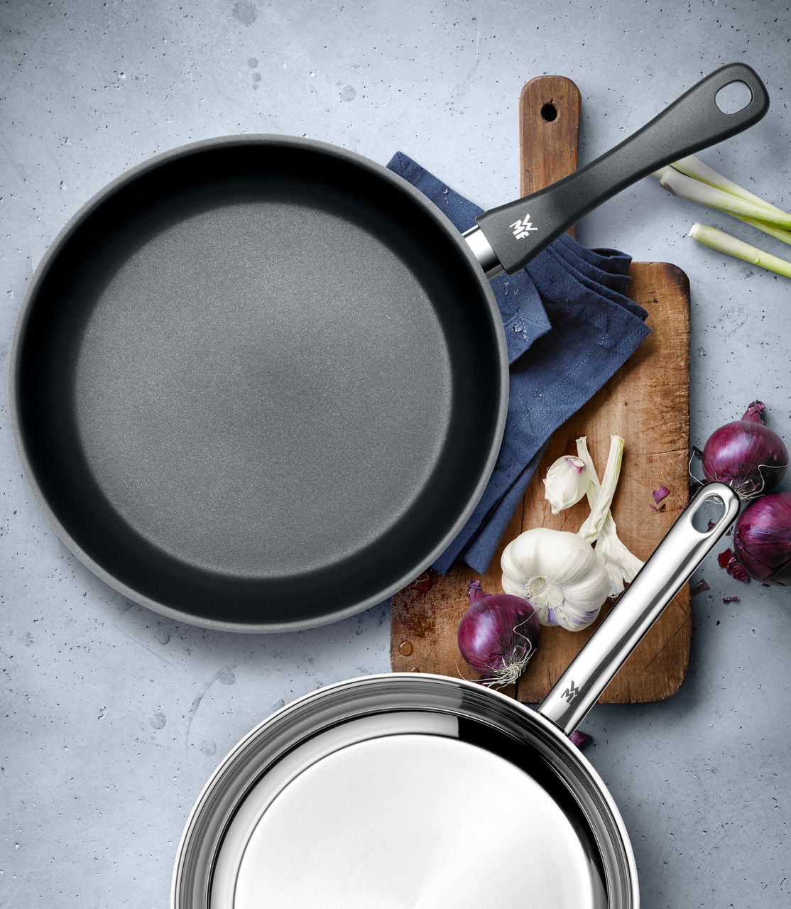 Zwei Pfannen-Set aus der Vogelperspektive, eine mit Antihaftbeschichtung und eine aus Edelstahl, auf einem Schneidebrett mit Zwiebeln und Knoblauch.