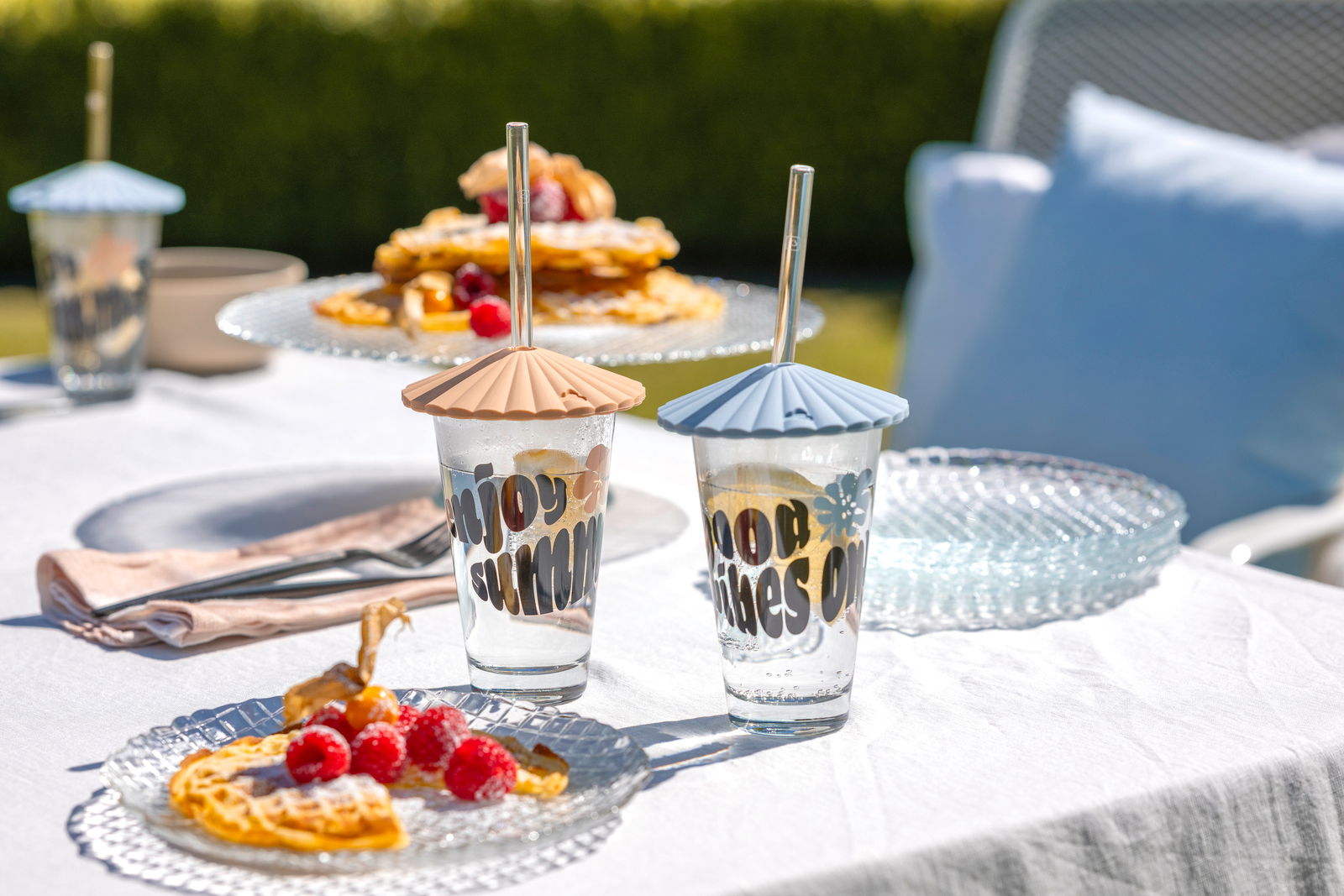 Zwei Trinkgläser mit bunten Deckeln und Strohhalmen auf einem gedeckten Tisch im Freien, umgeben von Waffeln und Himbeeren, aus der Perspektive von vorne.