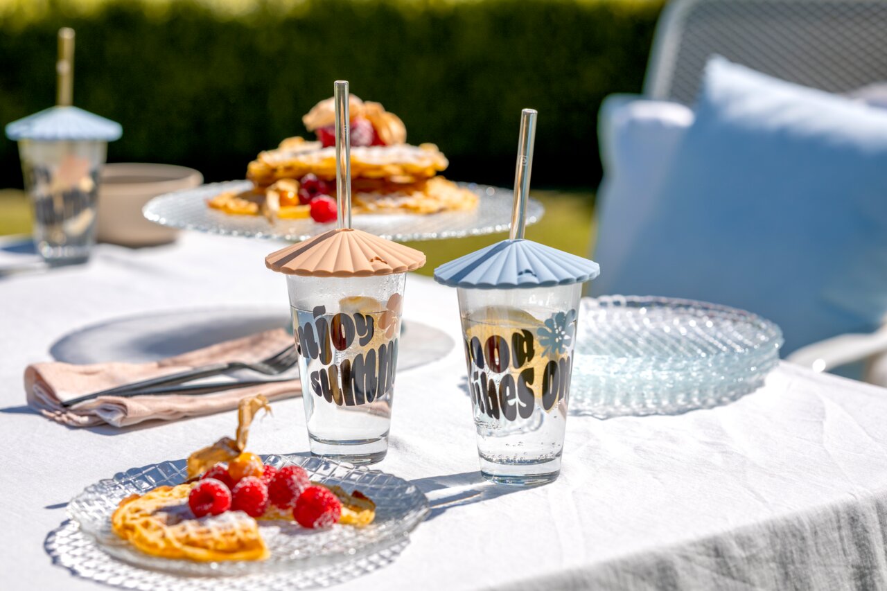 Twee drinkglazen met kleurrijke deksels en rietjes op een gedekte tafel in de open lucht, omringd door wafels en frambozen, gezien van voren.