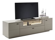Modern tv-lowboard in lichtgrijs met lades, houten accenten en een verlicht open vak, gefotografeerd vanaf de voorkant in een licht schuin perspectief; erop staan een flatscreen-tv en decoratie met kandelaars en boeken.