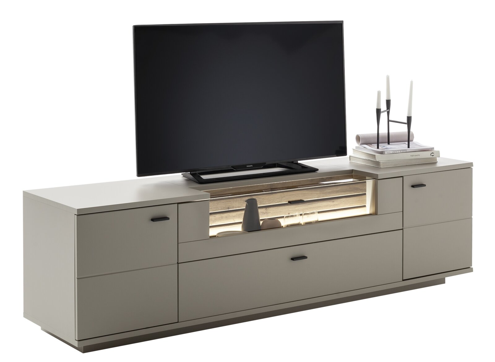 Modern tv-lowboard in lichtgrijs met lades, houten accenten en een verlicht open vak, gefotografeerd vanaf de voorkant in een licht schuin perspectief; erop staan een flatscreen-tv en decoratie met kandelaars en boeken.