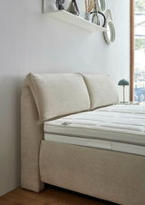 Seitliche Perspektive eines beigen Polsterbetts mit gepolstertem Kopfteil und weißer Matratze, im Hintergrund ein Fenster und eine Wand mit Dekoration.