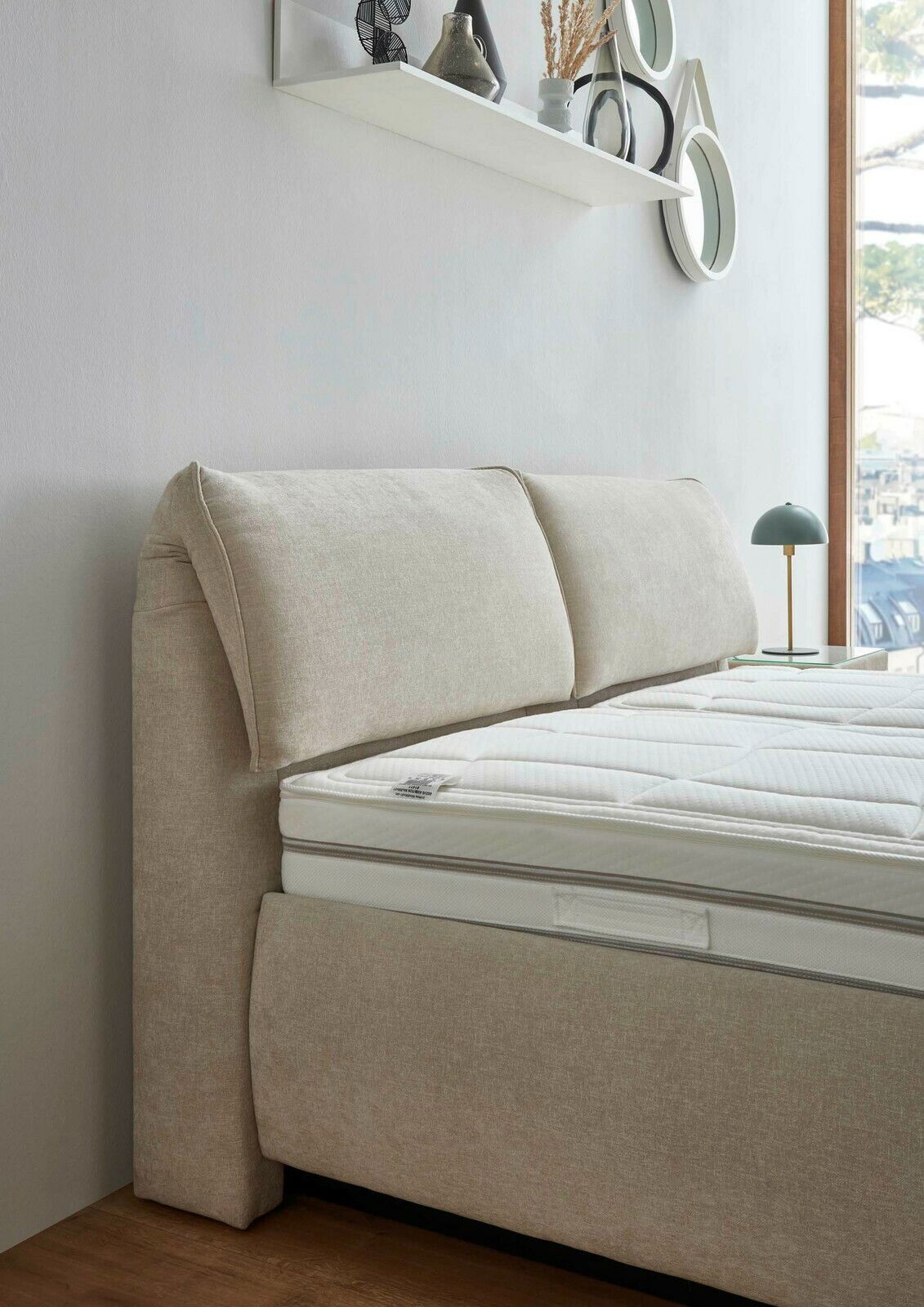 Seitliche Perspektive eines beigen Polsterbetts mit gepolstertem Kopfteil und weißer Matratze, im Hintergrund ein Fenster und eine Wand mit Dekoration.