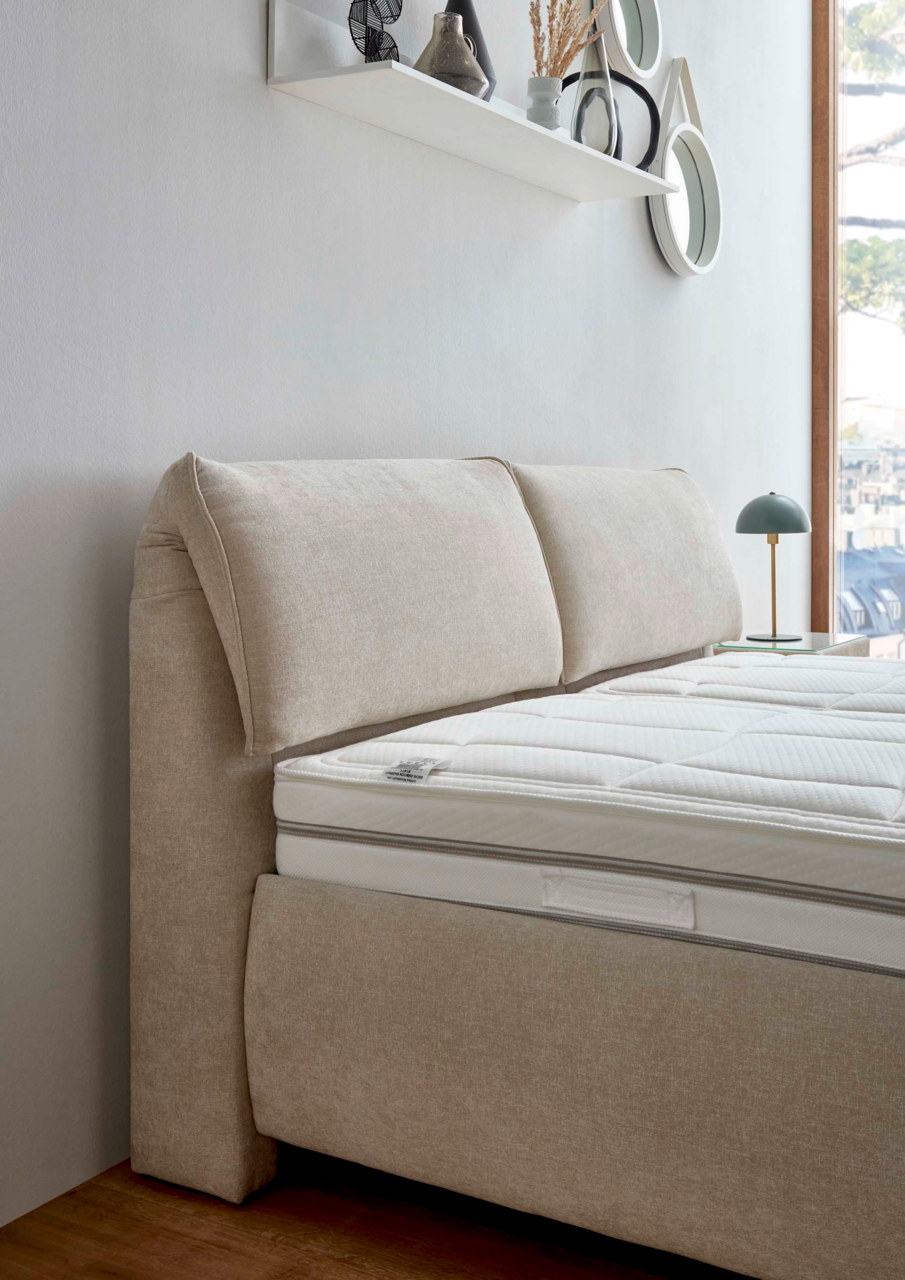 Seitliche Perspektive eines beigen Polsterbetts mit gepolstertem Kopfteil und weißer Matratze, im Hintergrund ein Fenster und eine Wand mit Dekoration.