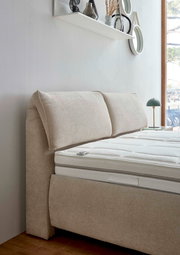 Seitliche Perspektive eines beigen Polsterbetts mit gepolstertem Kopfteil und weißer Matratze, im Hintergrund ein Fenster und eine Wand mit Dekoration.