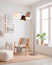 Moderne metalen boogvloerlamp met drie stoffen kappen in wit, zwart en grijs, brandend naast een houten fauteuil in een lichte woonkamer, gezien schuin van voren.
