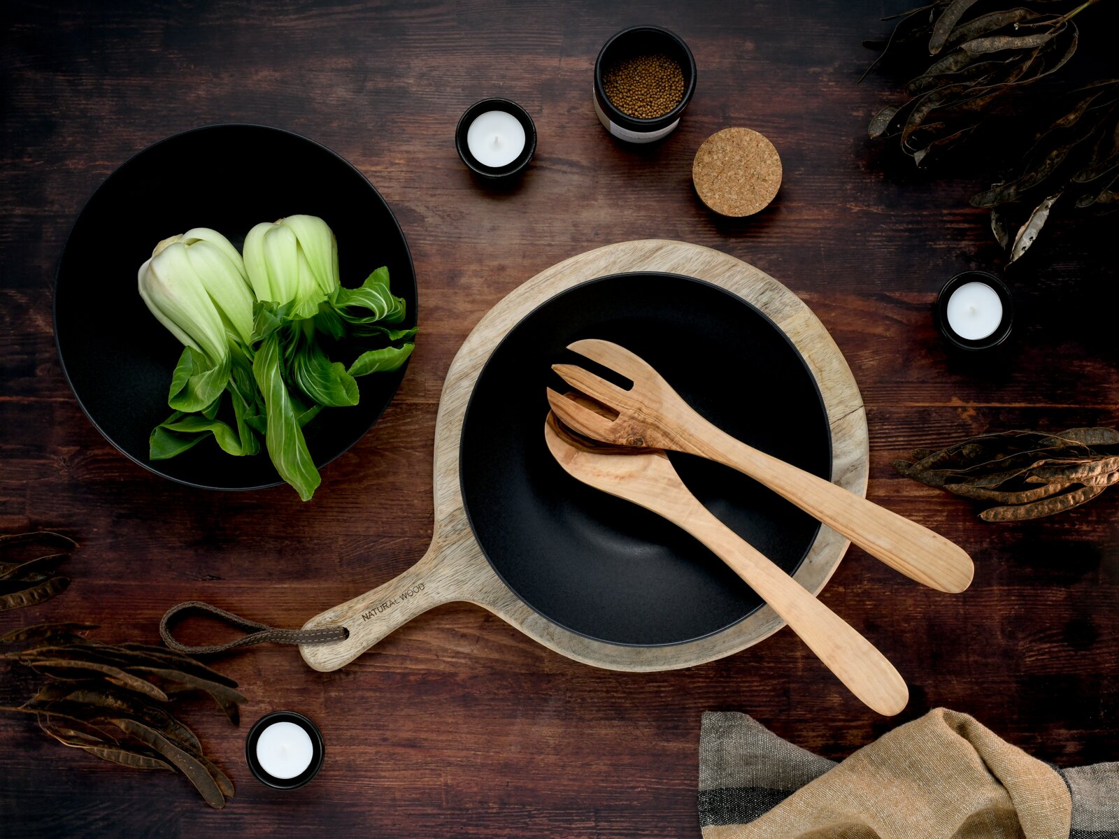 Draufsicht auf einen gedeckten Holztisch mit schwarzer Schale auf Holzbrett und Holz-Salatbesteck, daneben eine schwarze Schüssel mit frischem Pak Choi sowie kleine Kerzen und Gewürzdose.