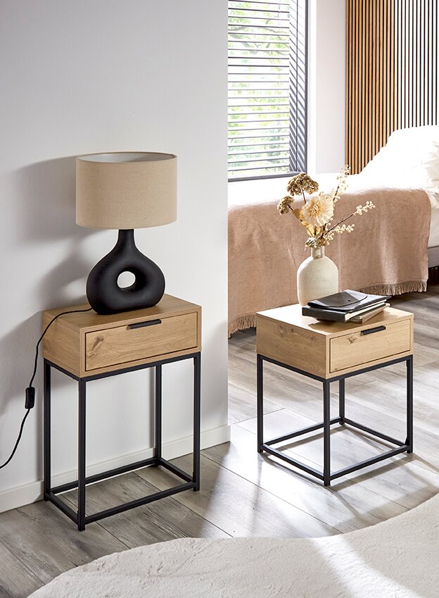 Zwei moderne Nachttische/Beistelltische aus hellem Holz mit Schublade und schwarzem Metallgestell im Schlafzimmer, fotografiert in leicht schräger Frontansicht auf Bodenhöhe; links mit Tischlampe, rechts mit Vase und Büchern.