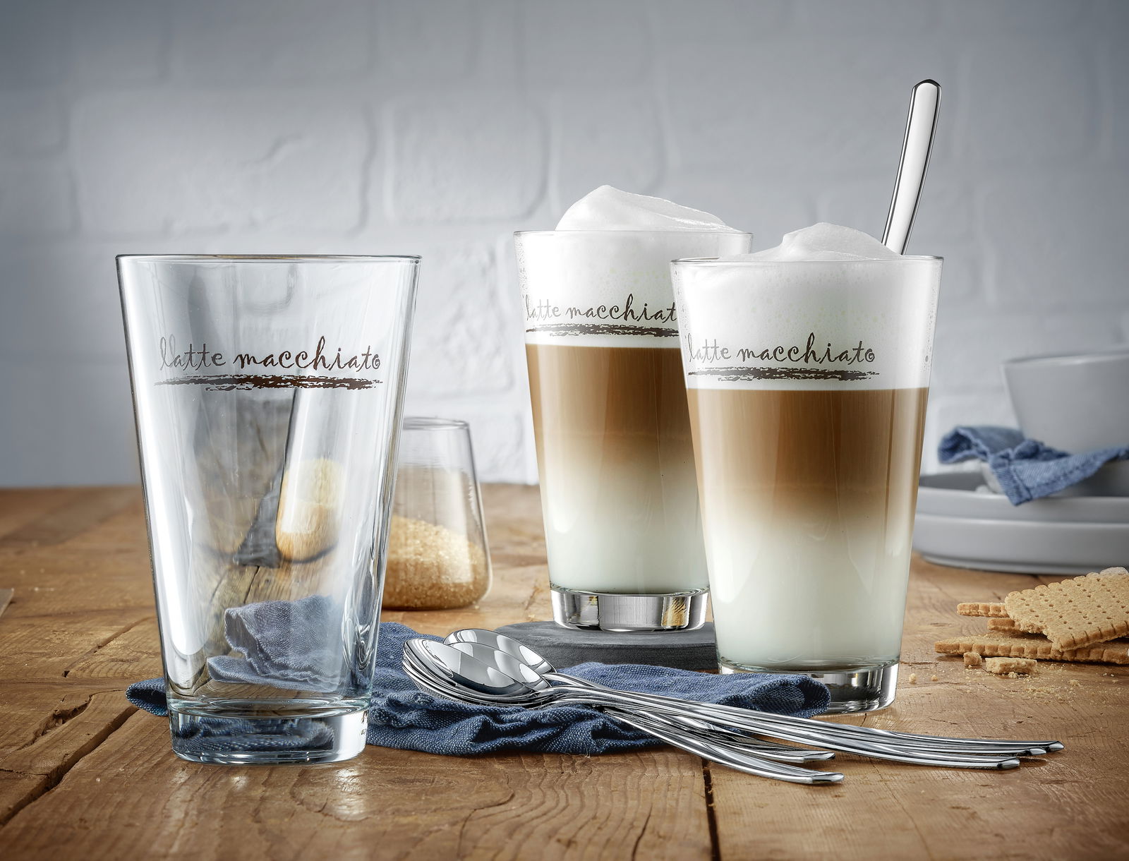 Latte Macchiato-Set mit zwei gefüllten Gläsern und einem leeren Glas, von vorne fotografiert, auf einem Holztisch mit Löffeln und Keksen.