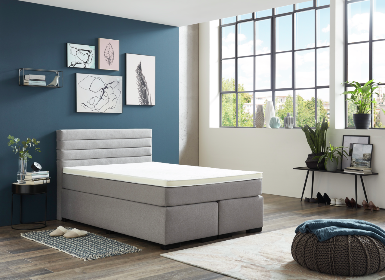 Modernes Boxspringbett in einem stilvoll eingerichteten Raum mit blauer Akzentwand und großen Fenstern, seitliche Perspektive.