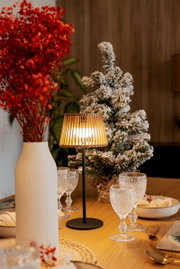 Stimmungsvolle Tischleuchte mit schwarzem Fuß und warm leuchtendem, lamellenförmigem Schirm auf einem gedeckten Holztisch, seitliche Perspektive im Wohnambiente mit Vase und winterlichem Deko-Baum im Hintergrund.