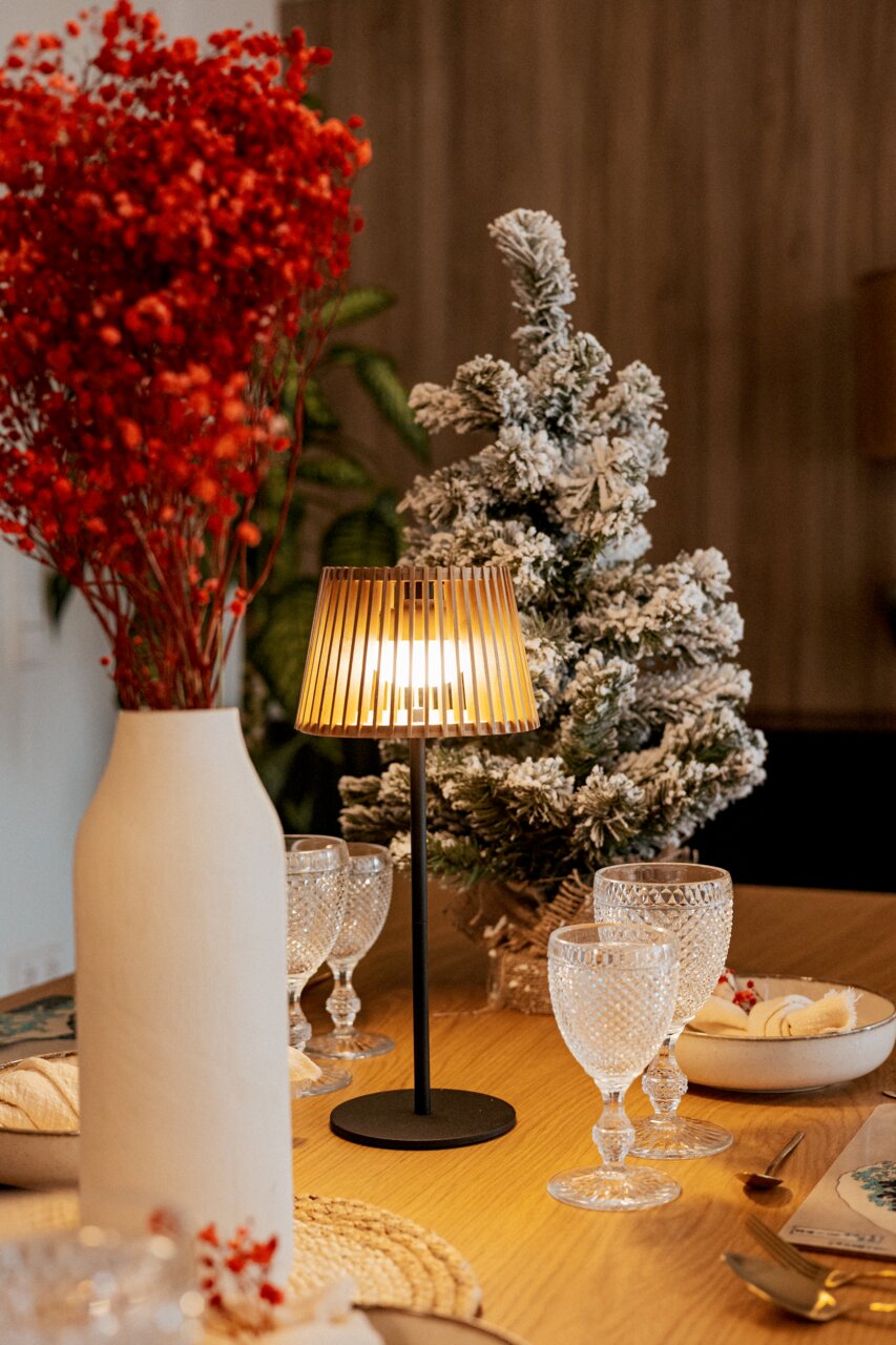 Stimmungsvolle Tischleuchte mit schwarzem Fuß und warm leuchtendem, lamellenförmigem Schirm auf einem gedeckten Holztisch, seitliche Perspektive im Wohnambiente mit Vase und winterlichem Deko-Baum im Hintergrund.