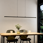 Moderne, slanke zwarte led-hanglamp in de vorm van een lange lichtbalk boven een houten tafel met twee stoelen, een vaas en serviesgoed, frontaal gefotografeerd vanuit een iets hoger perspectief