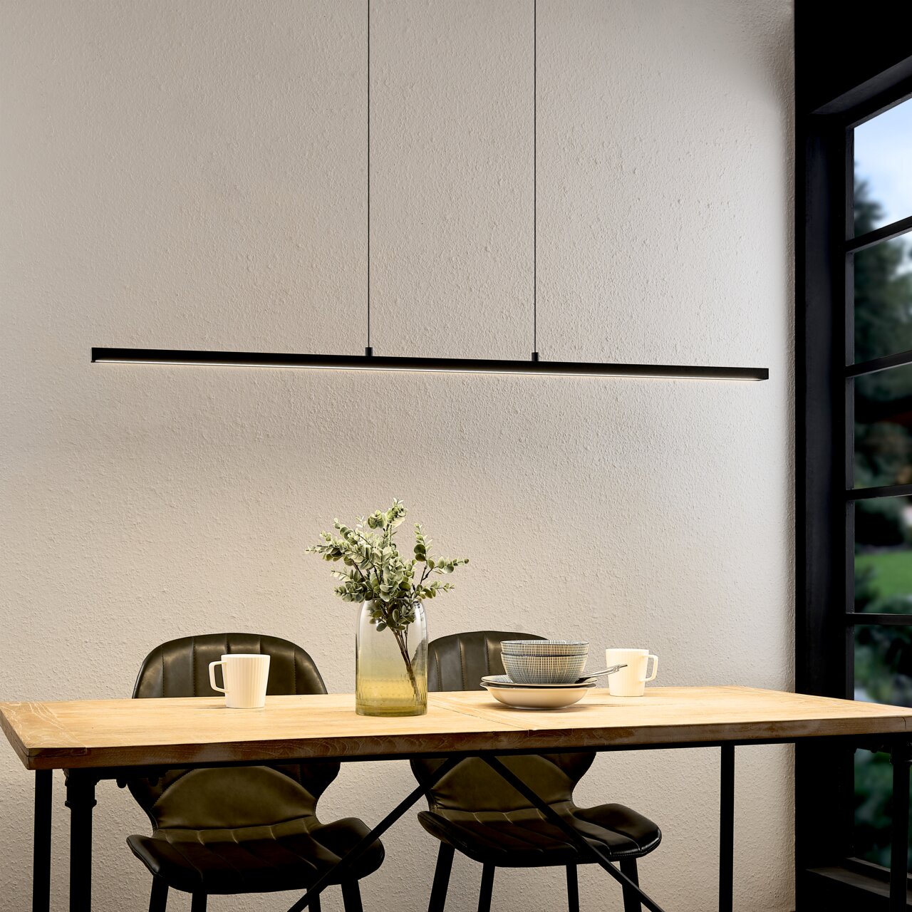 Moderne, slanke zwarte led-hanglamp in de vorm van een lange lichtbalk boven een houten tafel met twee stoelen, een vaas en serviesgoed, frontaal gefotografeerd vanuit een iets hoger perspectief