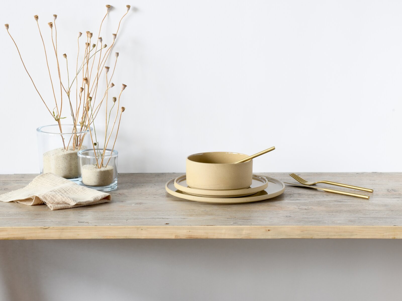 Beiges Keramik-Schüssel- und Teller-Set mit goldfarbenem Löffel sowie goldfarbenem Besteck auf Holztisch, seitliche Perspektive auf Tischhöhe vor weißem Hintergrund