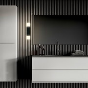 Moderne Badezimmer-Wandleuchte in Schwarz mit zylindrischem Opalglas, montiert neben einem Spiegel über einem Waschtisch, frontal aus leicht seitlicher Perspektive aufgenommen.