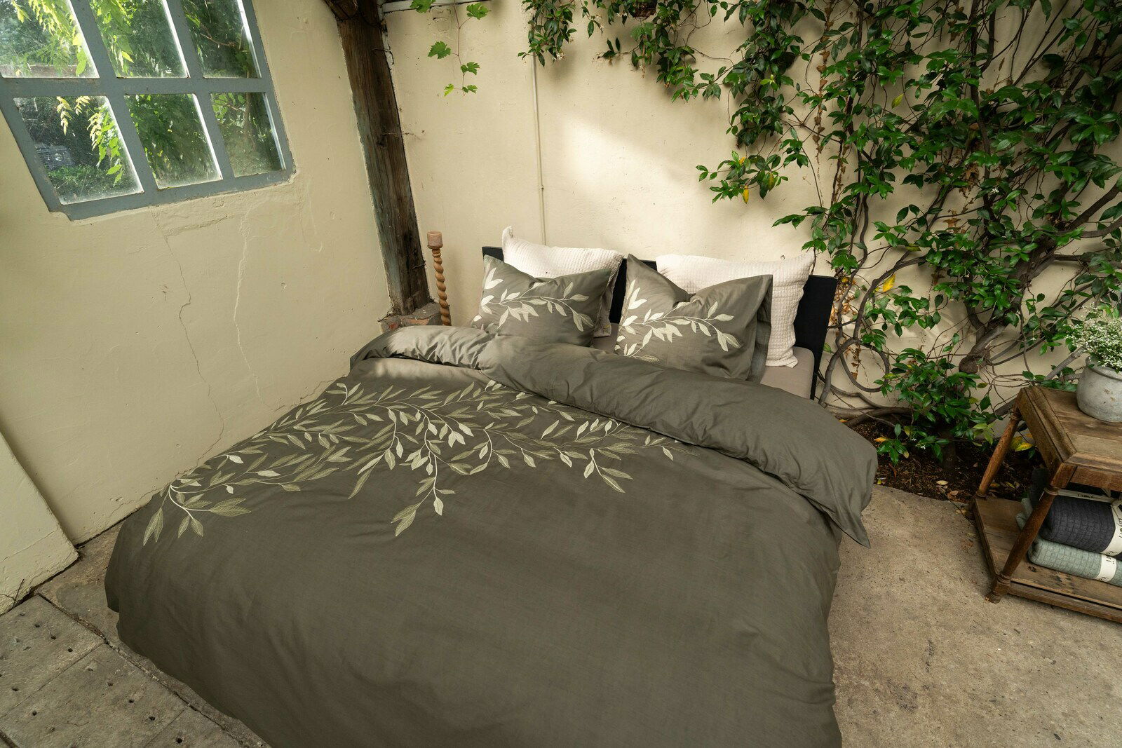 Bettwäsche-Set in einem natürlichen, grünen Farbton mit Blattmuster, auf einem Bett in einem rustikalen Raum mit Pflanzen im Hintergrund, aus der Vogelperspektive fotografiert.