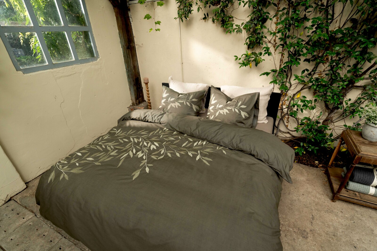 Bettwäsche-Set in einem natürlichen, grünen Farbton mit Blattmuster, auf einem Bett in einem rustikalen Raum mit Pflanzen im Hintergrund, aus der Vogelperspektive fotografiert.