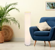 Schlanke, zylindrische Stehlampe mit weißem Stoffschirm und Metallfuß neben blauem Sessel in modernem Wohnzimmer, frontale Perspektive.