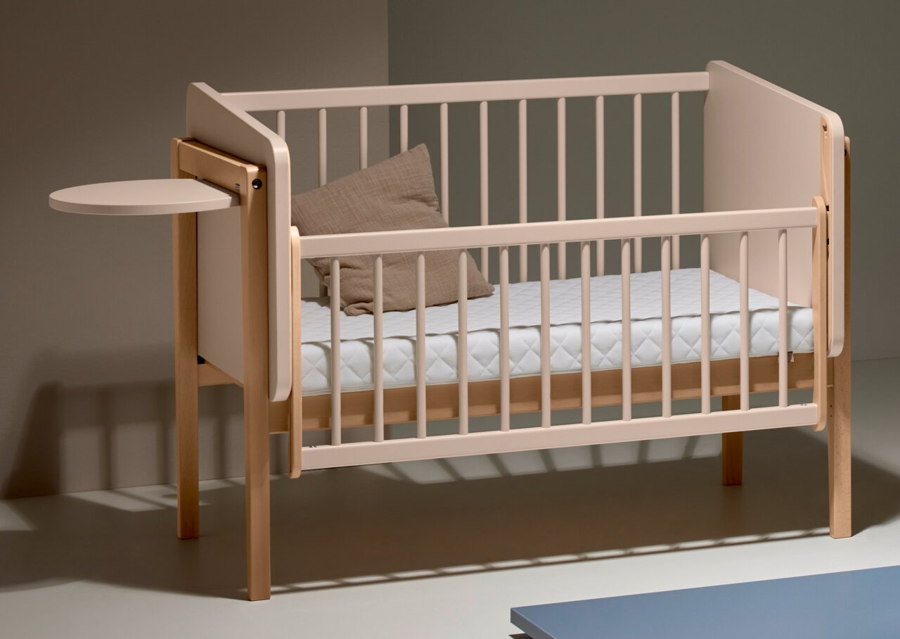 Kinderbett mit hellem Holzrahmen und abgerundeten Ecken, seitlich angebrachtem kleinen Tischchen, von schräg vorne fotografiert.
