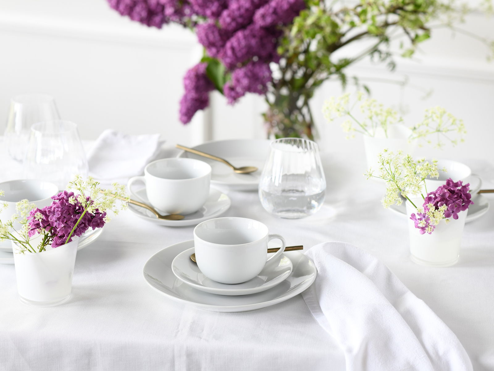 Elegantes Kaffeeservice 18-tlg auf einem gedeckten Tisch mit weißen Tassen und Tellern, goldenen Löffeln und dekorativen lila und weißen Blumen, seitliche Perspektive.