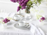 Elegantes Kaffeeservice 18-tlg auf einem gedeckten Tisch mit weißen Tassen und Tellern, goldenen Löffeln und dekorativen lila und weißen Blumen, seitliche Perspektive.