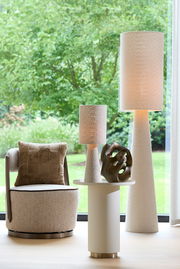 Ein modernes Wohnzimmer mit einem beigen Sessel, einem runden Beistelltisch und zwei hohen, strukturierten Stehlampen. Im Vordergrund steht eine abstrakte Skulptur auf dem Tisch. Die Perspektive zeigt das Arrangement von vorne mit einem Blick auf den grün