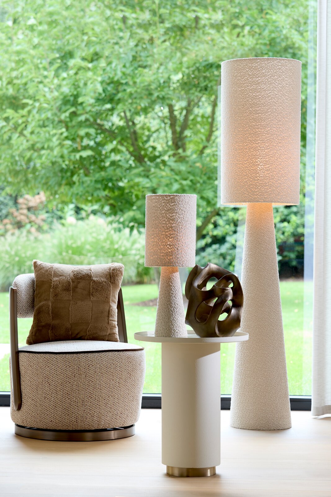 Een moderne woonkamer met een beige fauteuil, een rond bijzettafeltje en twee hoge, gestructureerde staande lampen. Op de voorgrond staat een abstract beeld op het tafeltje. Het perspectief toont de opstelling van voren, met uitzicht op de groene tuin op 