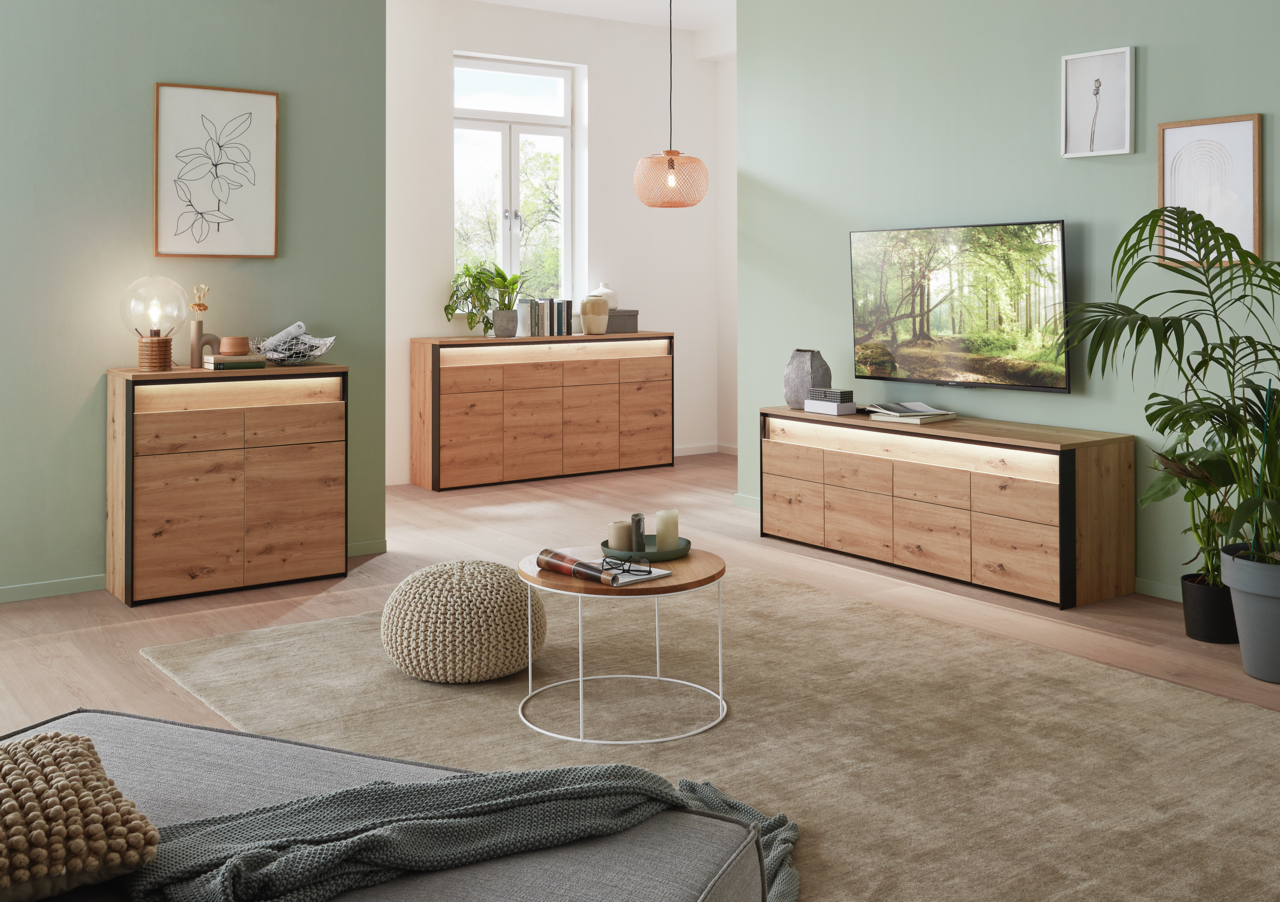 Moderne Wohnzimmermöbel aus Holz mit Sideboard, Kommode und TV-Lowboard in einem hellen Raum mit grüner Wand und dekorativen Pflanzen, seitliche Perspektive.