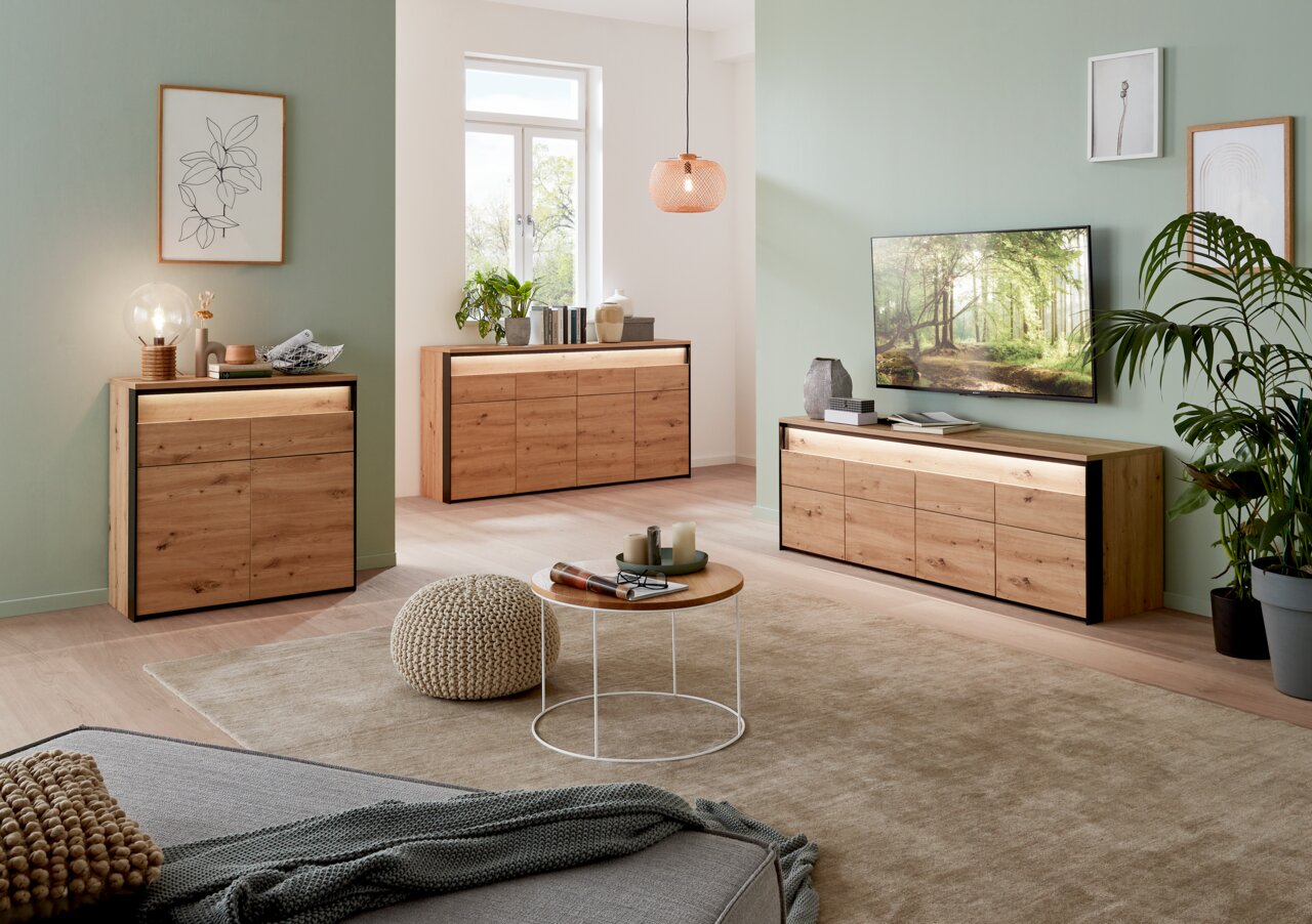 Voleo Sideboard Moderne Wohnzimmermöbel aus Holz mit Sideboard, Kommode und TV-Lowboard in einem hellen Raum mit grüner Wand und dekorativen Pflanzen, seitliche Perspektive.