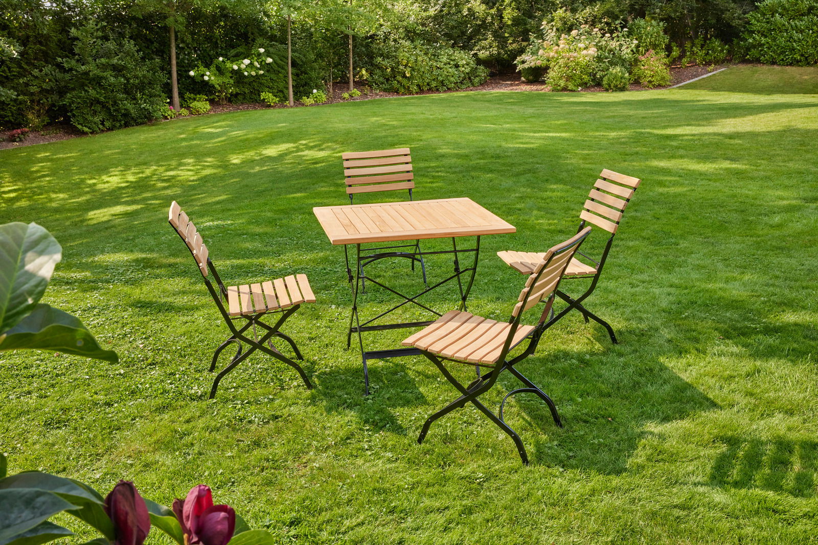 Kleiner, klappbarer Gartentisch mit vier passenden Stühlen auf einer grünen Rasenfläche, umgeben von Bäumen und Sträuchern, aus der Vogelperspektive fotografiert.
