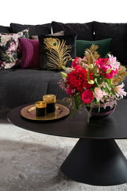 Großes Sofa mit dekorativen Kissen in verschiedenen Farben und Mustern, im Vordergrund ein runder Couchtisch mit einem Blumenarrangement und goldenen Kerzenhaltern, seitliche Perspektive.