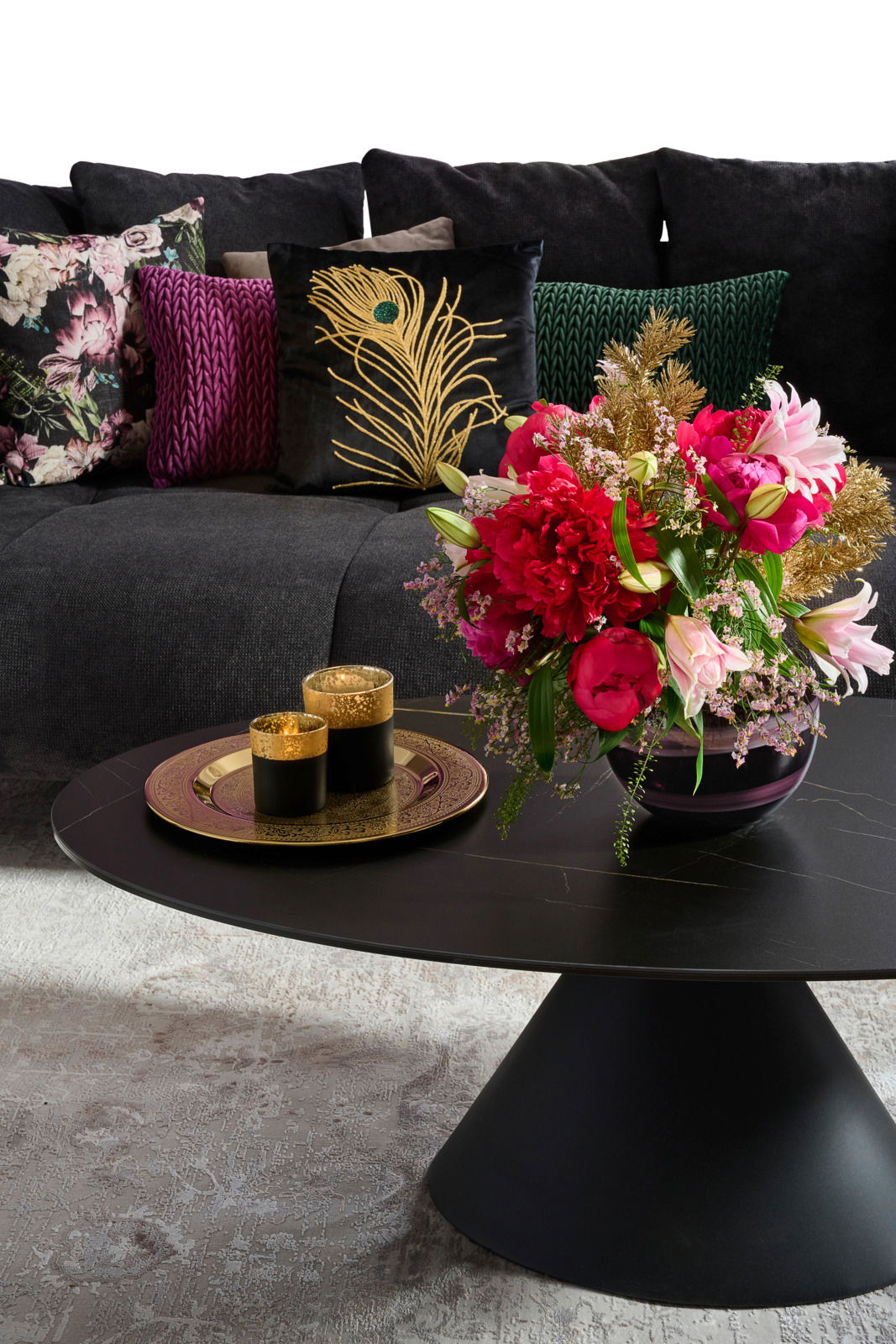Großes Sofa mit dekorativen Kissen in verschiedenen Farben und Mustern, im Vordergrund ein runder Couchtisch mit einem Blumenarrangement und goldenen Kerzenhaltern, seitliche Perspektive.