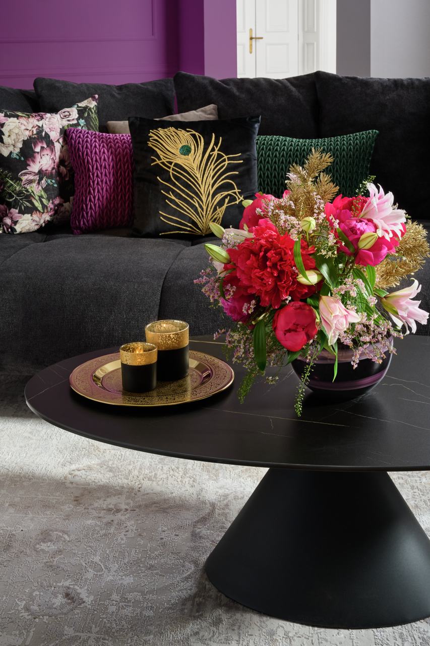 Großes Sofa mit dekorativen Kissen in verschiedenen Farben und Mustern, im Vordergrund ein runder Couchtisch mit einem Blumenarrangement und goldenen Kerzenhaltern, seitliche Perspektive.