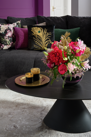 Großes Sofa mit dekorativen Kissen in verschiedenen Farben und Mustern, im Vordergrund ein runder Couchtisch mit einem Blumenarrangement und goldenen Kerzenhaltern, seitliche Perspektive.