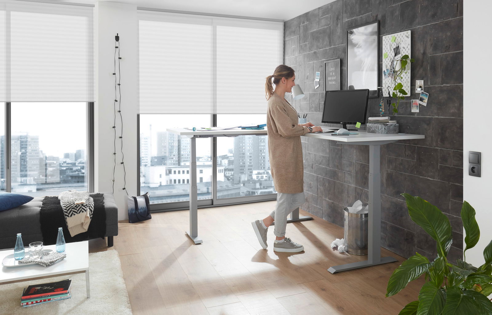 Frau arbeitet an einem höhenverstellbaren Eckschreibtisch in einem modernen Büro mit Stadtblick, seitliche Perspektive.