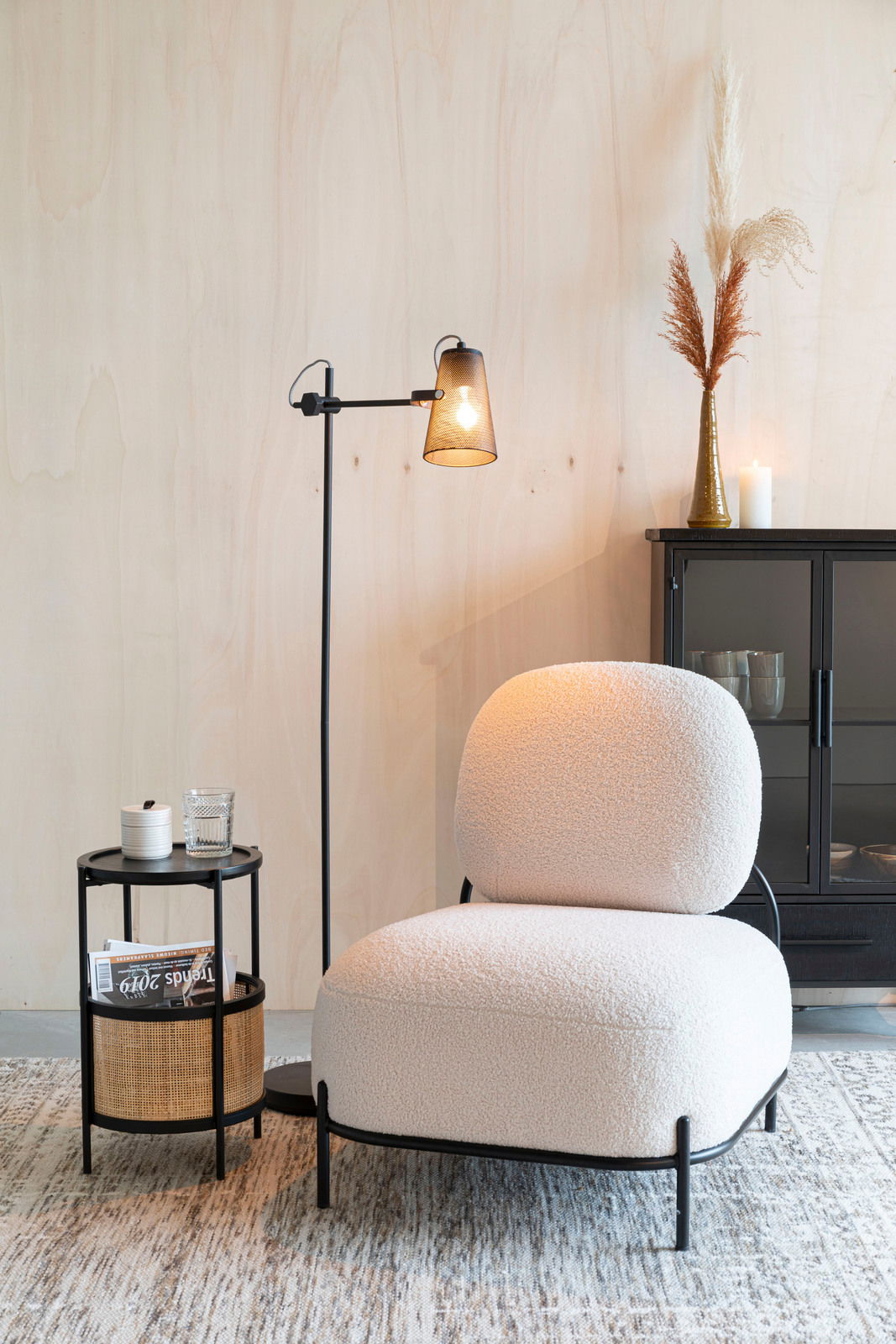 Beistelltisch MAKATO aus schwarzem Metall mit zwei Ebenen, eine mit Rattan-Elementen, im modernen Wohnzimmer neben einem Sessel, aus seitlicher Perspektive.