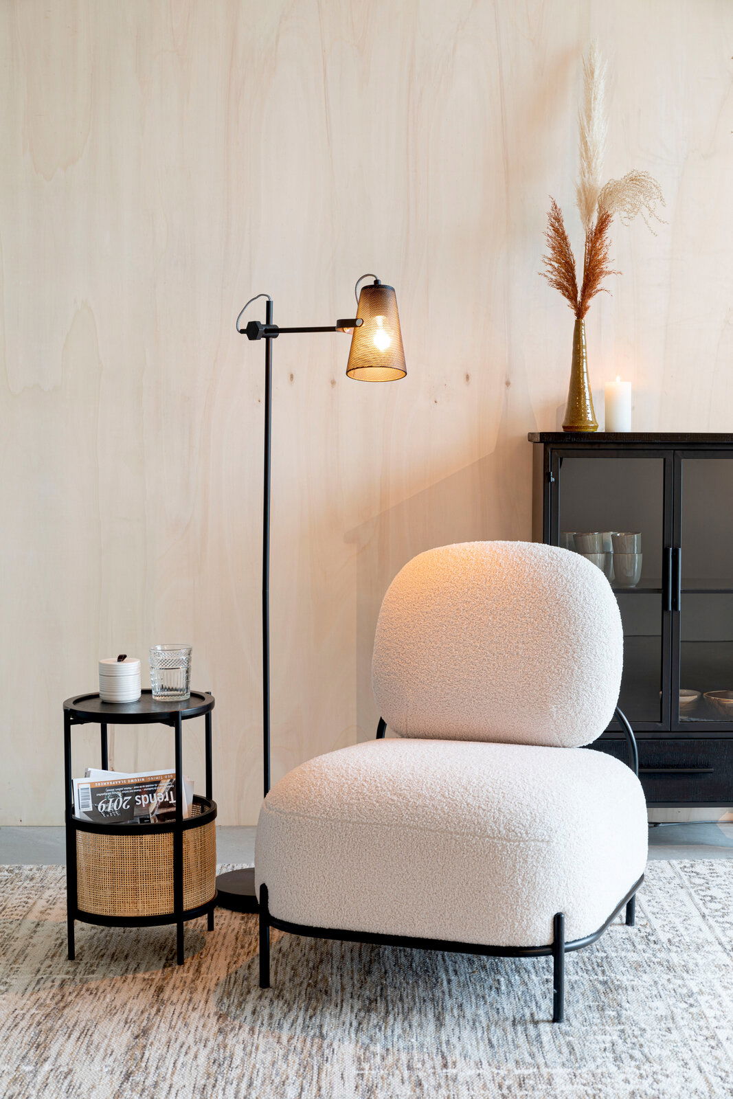 Beistelltisch MAKATO aus schwarzem Metall mit zwei Ebenen, eine mit Rattan-Elementen, im modernen Wohnzimmer neben einem Sessel, aus seitlicher Perspektive.