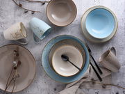 Vogelperspektive auf ein stilvolles 16-teiliges Kombiservice-Set mit Tellern, Schalen und Tassen in sanften Beige- und Blautönen, dekoriert mit Zweigen und Besteck.