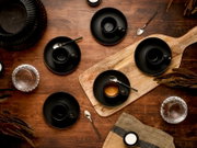 Draufsicht auf mehrere schwarze Espressotassen mit Untertassen und Löffeln auf einem rustikalen Holztisch, teils auf einem Holzbrett arrangiert, mit Wassergläsern und Deko-Elementen.
