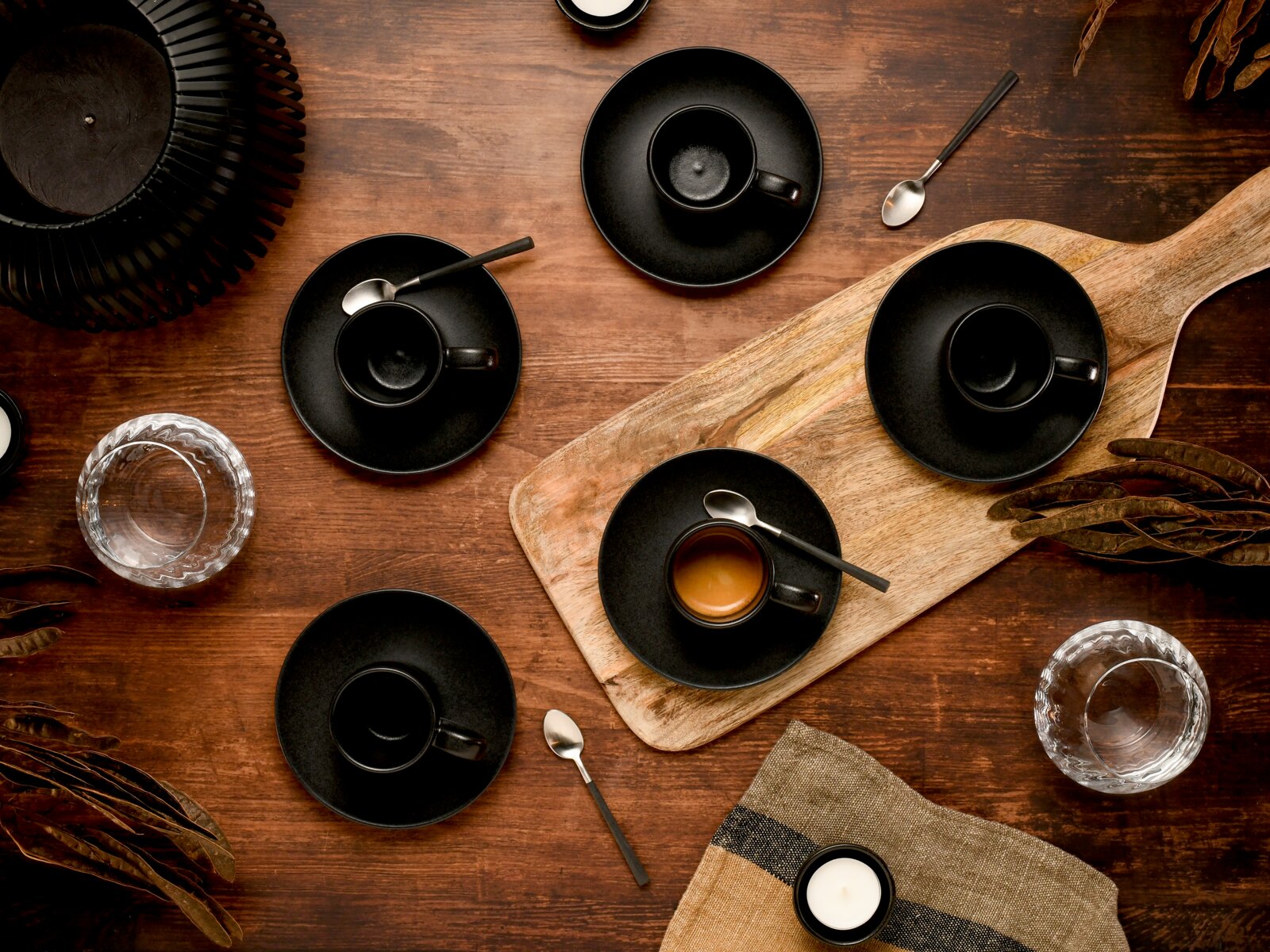 Draufsicht auf mehrere schwarze Espressotassen mit Untertassen und Löffeln auf einem rustikalen Holztisch, teils auf einem Holzbrett arrangiert, mit Wassergläsern und Deko-Elementen.