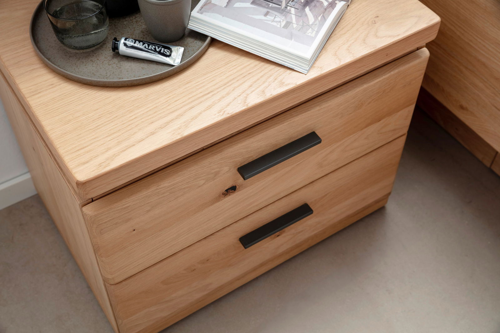 Nachtkonsole aus Holz mit zwei Schubladen, schwarze Griffe, von oben fotografiert, dekoriert mit einem Tablett, einer Zahnpastatube und einem Buch.