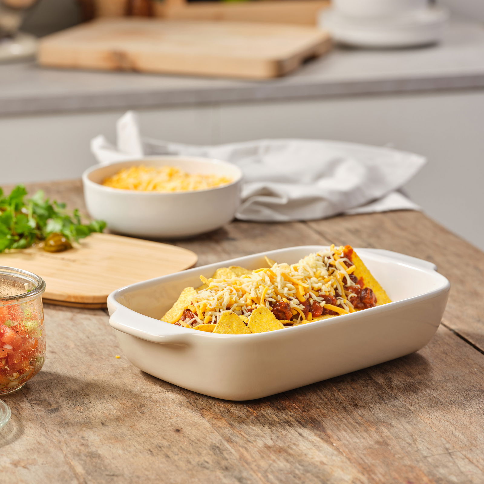 Keramik-Auflaufform mit Deckel, gefüllt mit überbackenen Nachos, auf einem Holztisch in einer Küchenumgebung, seitliche Perspektive
