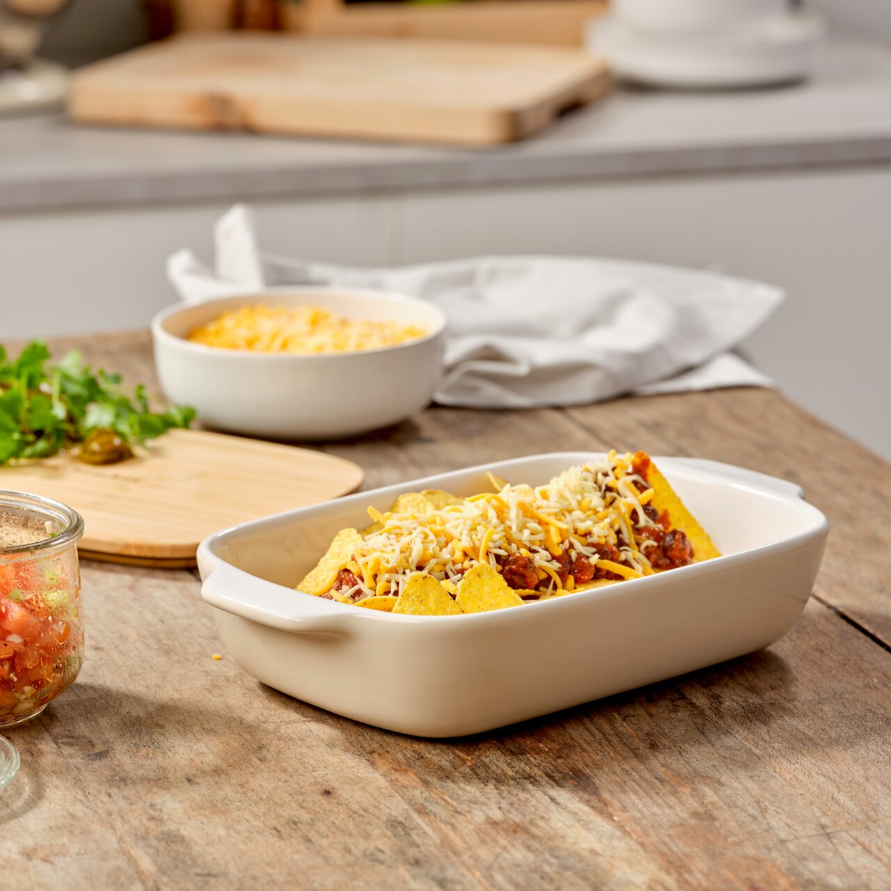 Keramik-Auflaufform mit Deckel, gefüllt mit überbackenen Nachos, auf einem Holztisch in einer Küchenumgebung, seitliche Perspektive