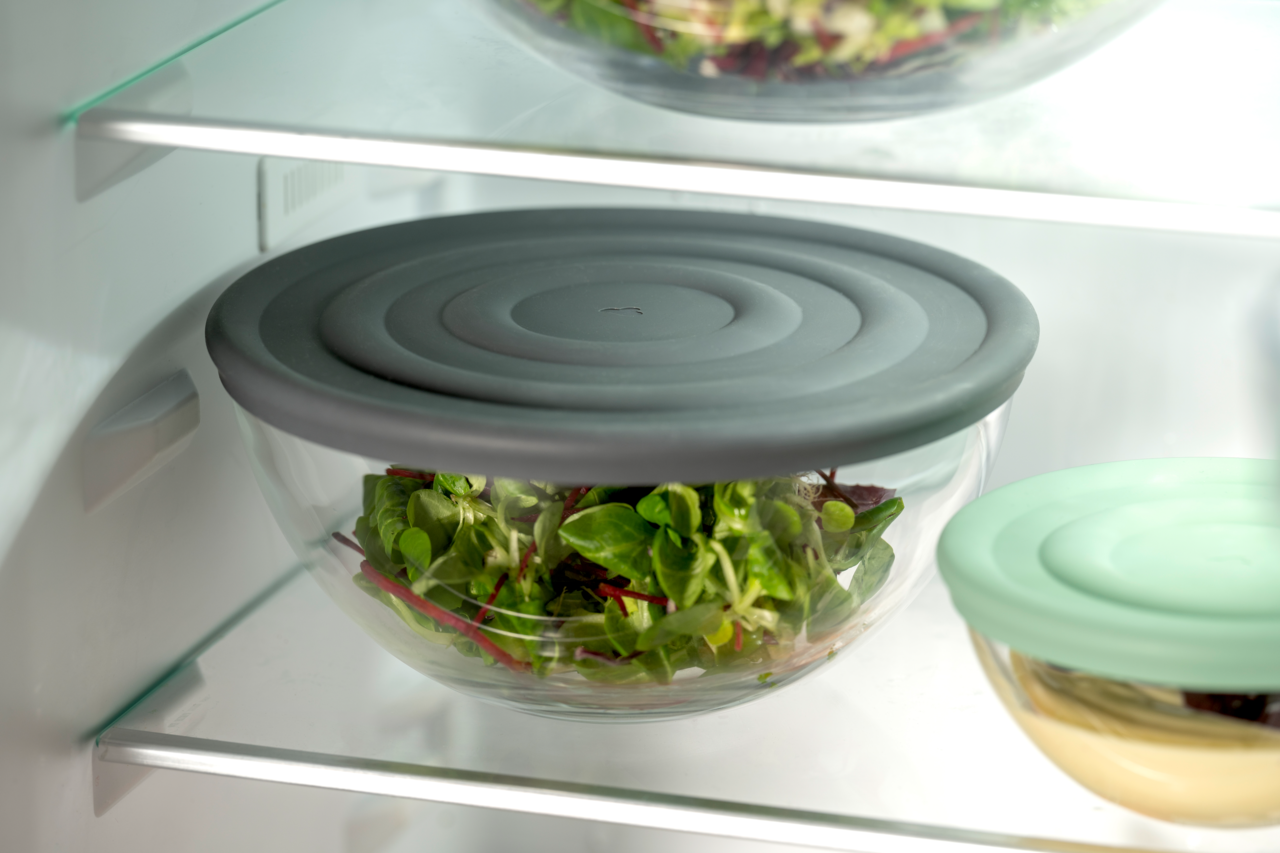 Glas-Schale mit grauem Deckel, gefüllt mit frischem Salat, im Kühlschrank von der Seite betrachtet.