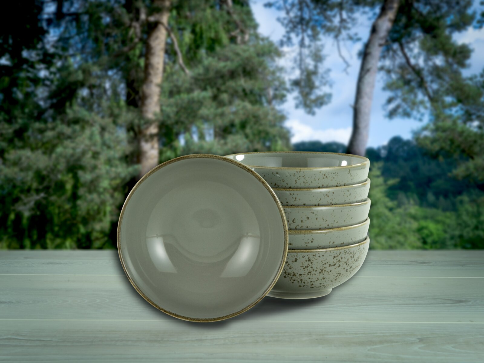 Stapel aus fünf gesprenkelten Keramikschalen in Creme/Beige mit braunem Rand auf Holzoberfläche vor unscharfem Waldhintergrund, eine Schale vorne frontal von oben leicht geneigt gezeigt