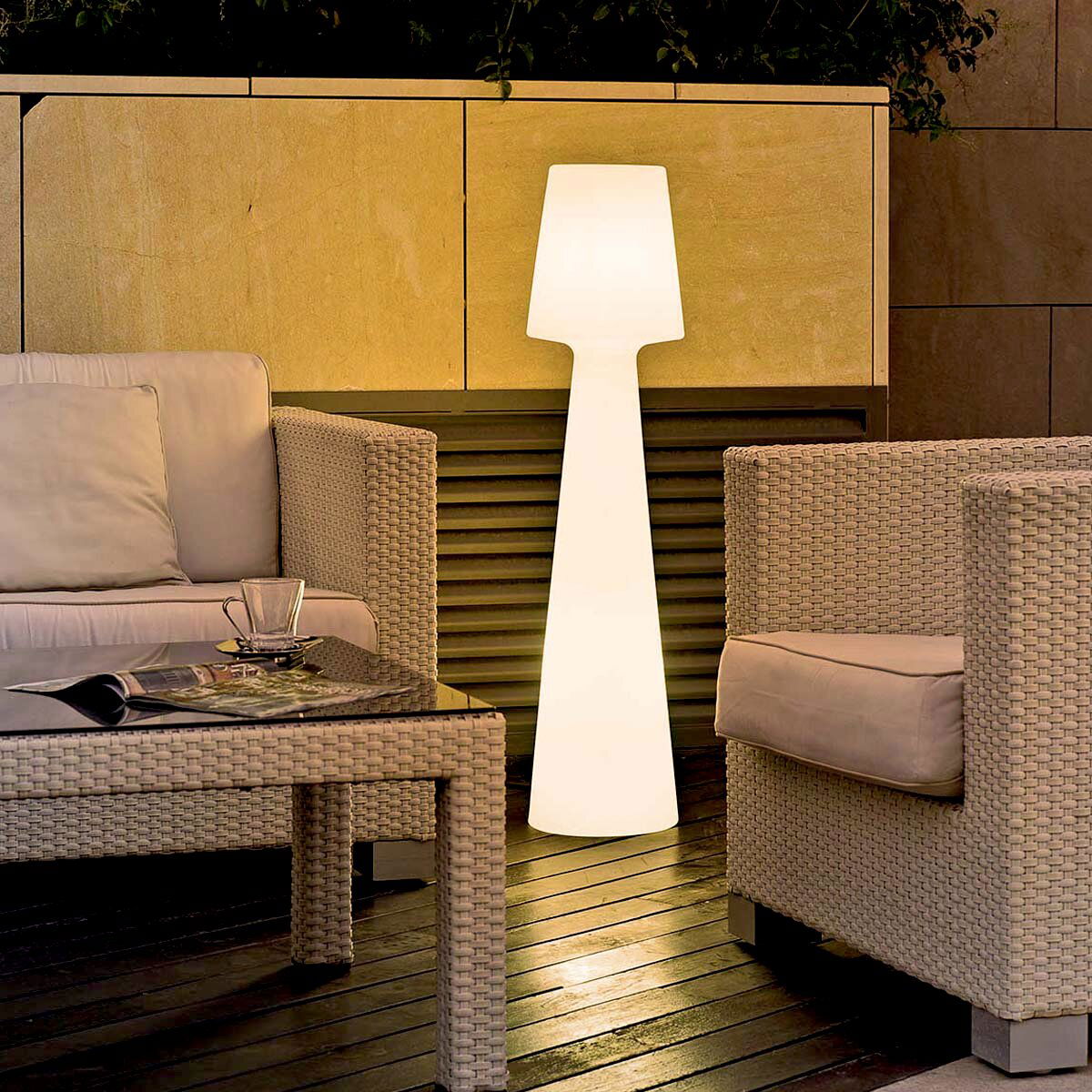 Leuchtende weiße Outdoor-Stehleuchte in modernem Design zwischen Rattan-Loungemöbeln auf einer Terrasse, frontale Perspektive auf das Produkt