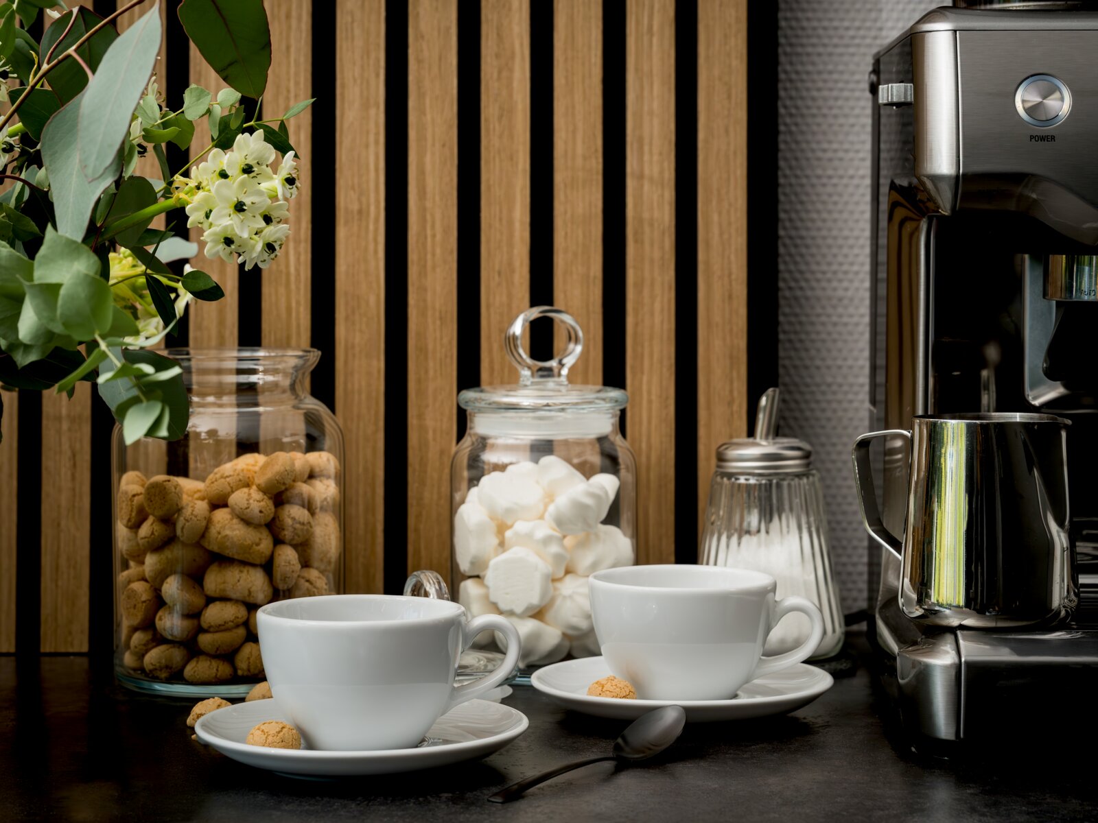 creaTable Kaffeetasse Set 8-tlg BISTROT Schwarze Kaffeemaschine mit Edelstahl-Milchkännchen neben zwei weißen Kaffeetassen und Vorratsgläsern mit Keksen und Marshmallows auf dunkler Arbeitsplatte, Frontalansicht mit leicht seitlicher Perspektive von links.