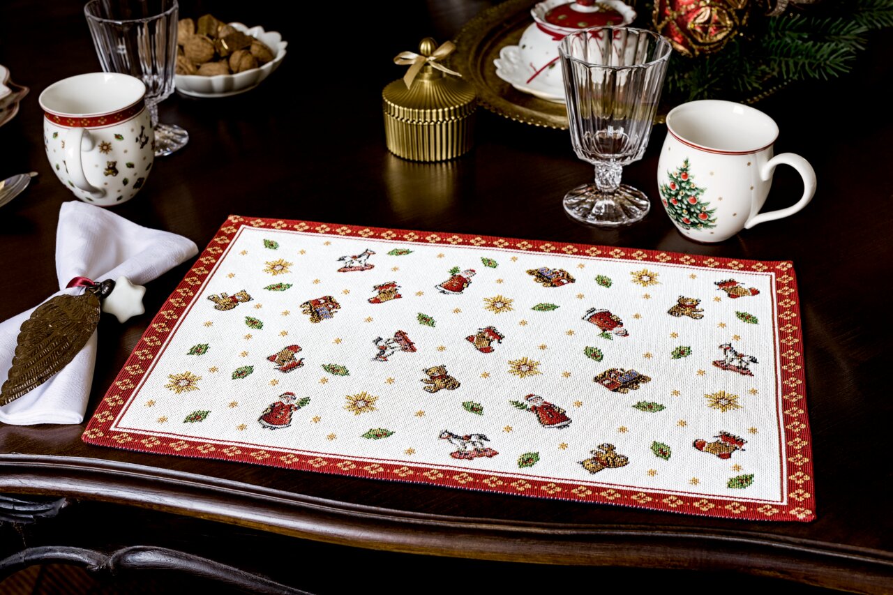 Kerstachtige gobelin-placemat met speelgoedmotieven en een rode rand, van bovenaf gefotografeerd op een gedekte tafel.