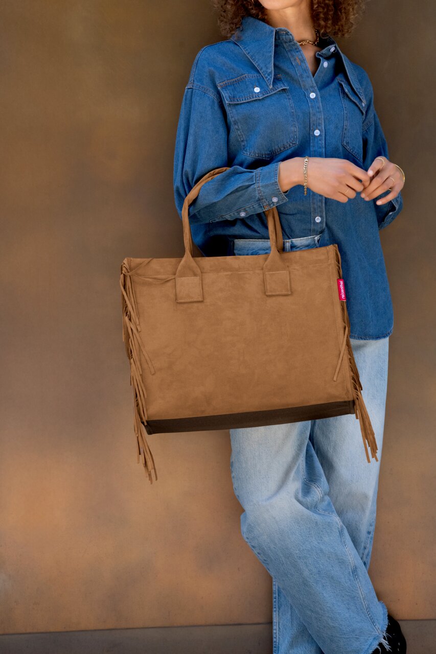 Braune Wildleder-Optik Shopper-Tasche mit langen Henkeln und Fransen an den Seiten, getragen von einer Frau im Jeanshemd und hellen Jeans vor braunem Hintergrund, Frontansicht in leichter Seitenperspektive.